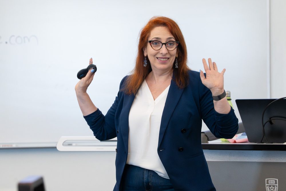 Ingrid en un aula, sonriendo, con las manos en alto y el control de la presentación